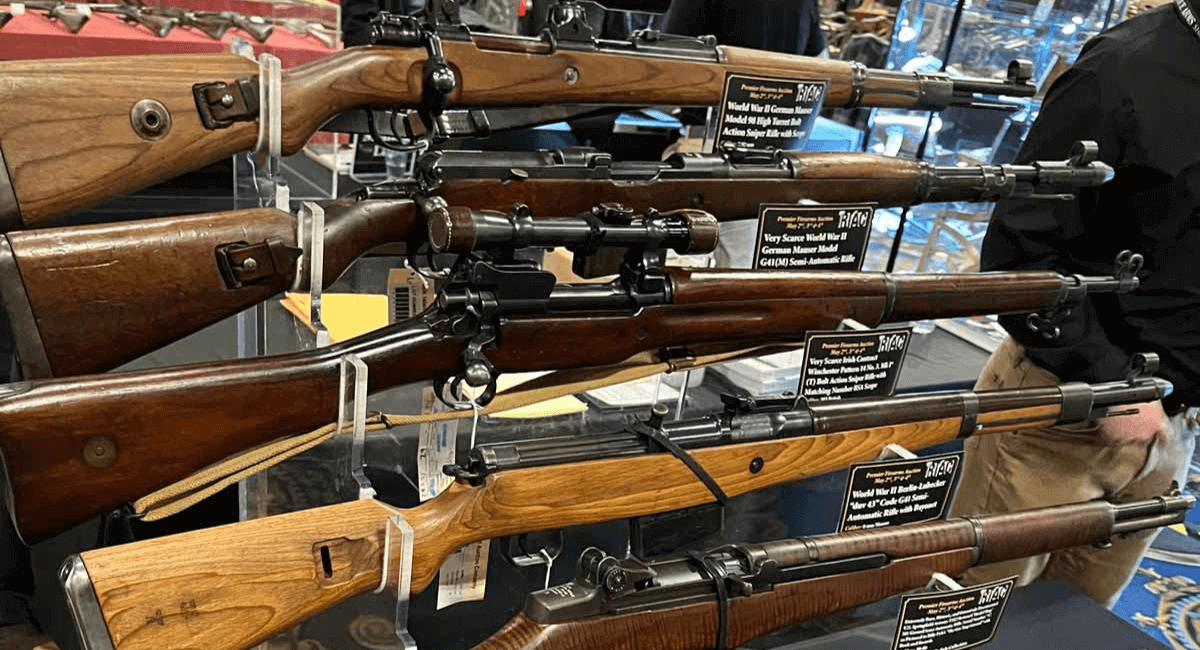 Four vintage bolt-action rifles are displayed horizontally on racks, each with a descriptive information card attached, at what appears to be a gun show or exhibition.