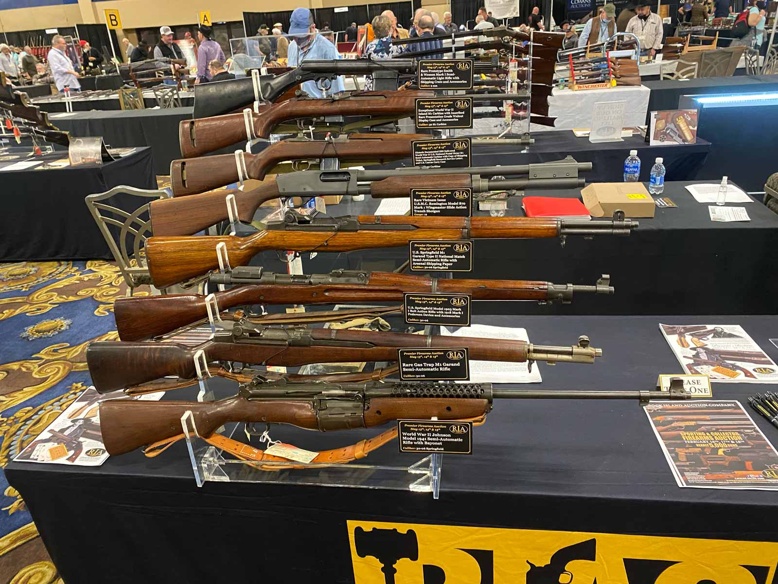 A display of vintage rifles arranged horizontally on a stand at a gun show, with informational placards and tables in the background.