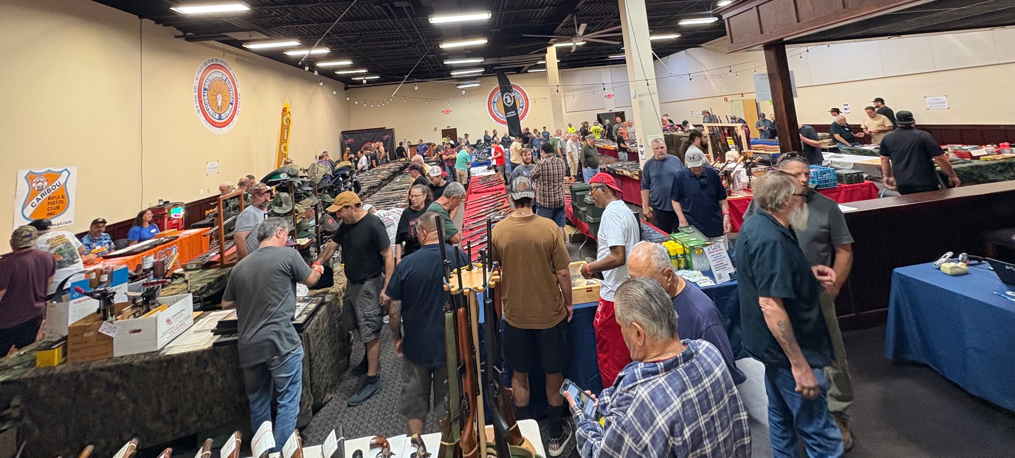 Large crowded indoor gun show with numerous tables displaying firearms and accessories; people are browsing, talking, and examining items under bright overhead lighting.
